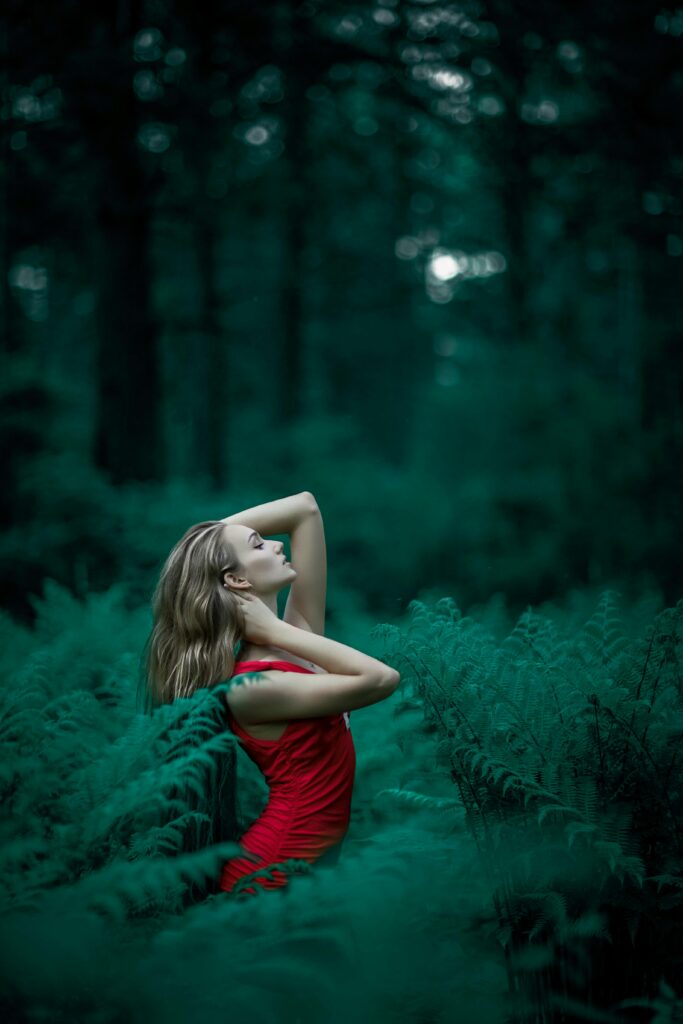 pexels-photo-1921168-1921168 A woman poses in a vibrant red dress amidst lush green ferns in an enchanting forest setting.