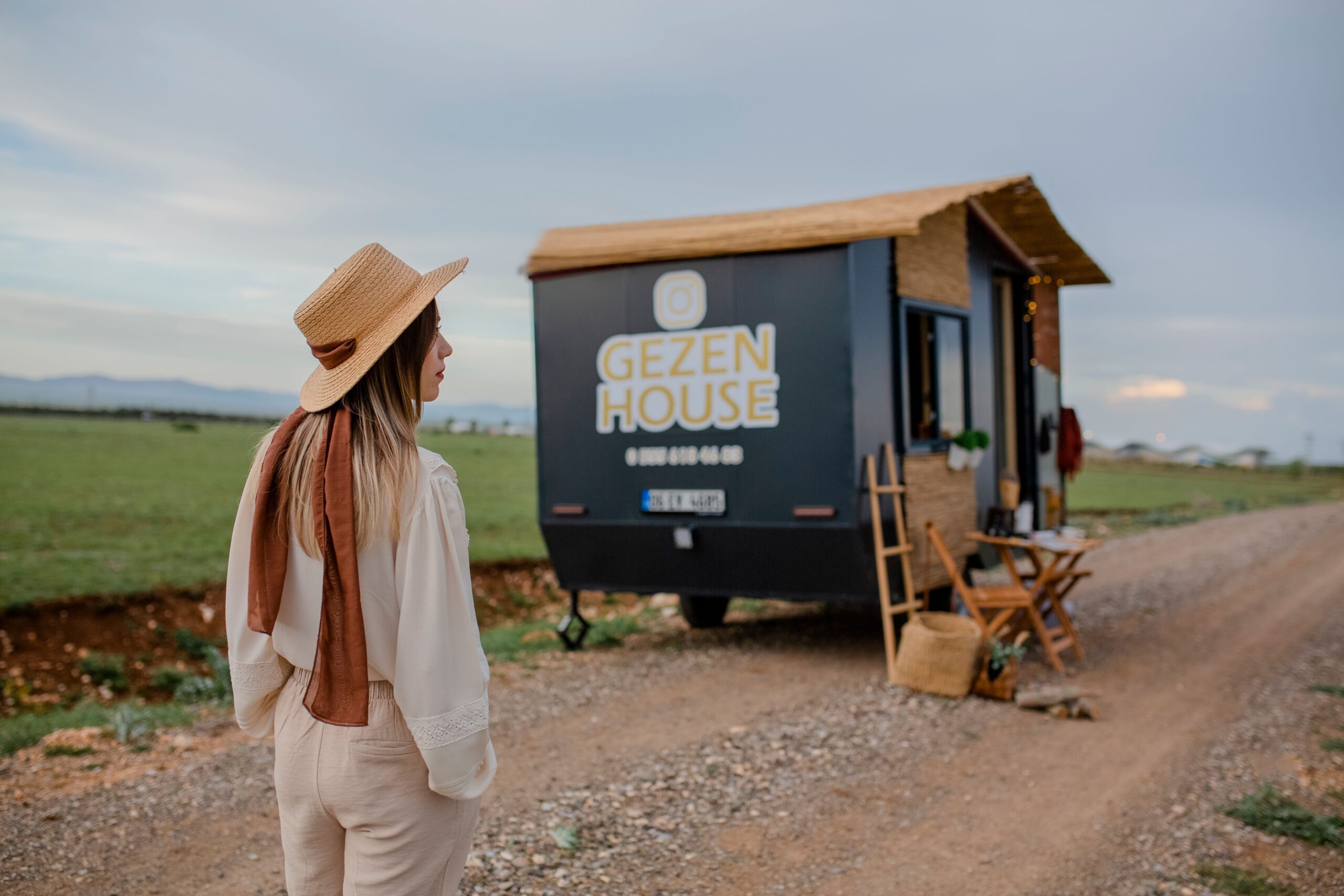A woman wearing a straw hat stands near a tiny house on a dirt road in a rural area, showcasing serenity and simplicity.