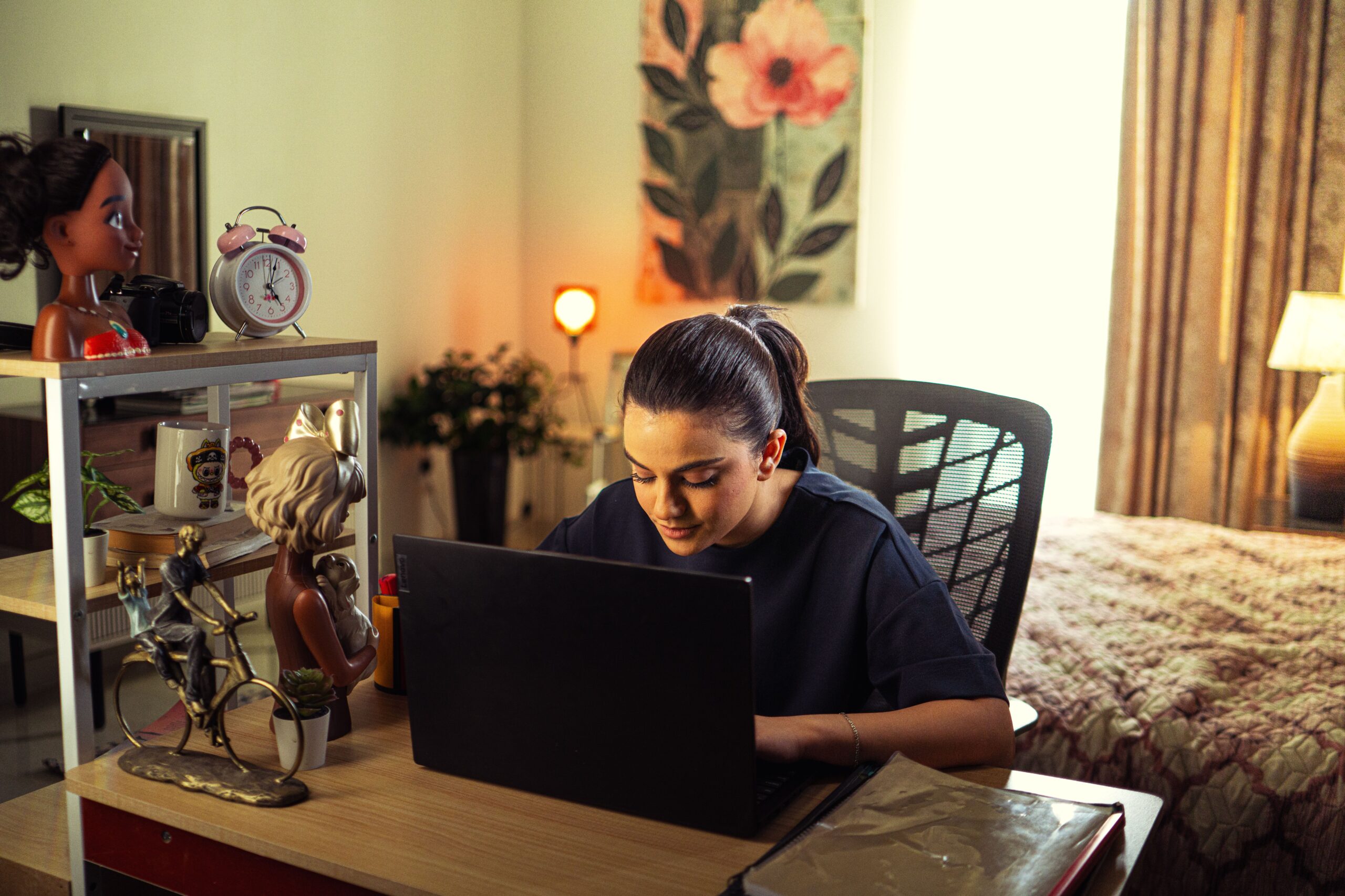 A woman focusing on her laptop in a cozy home office surrounded by decor and warm lighting.