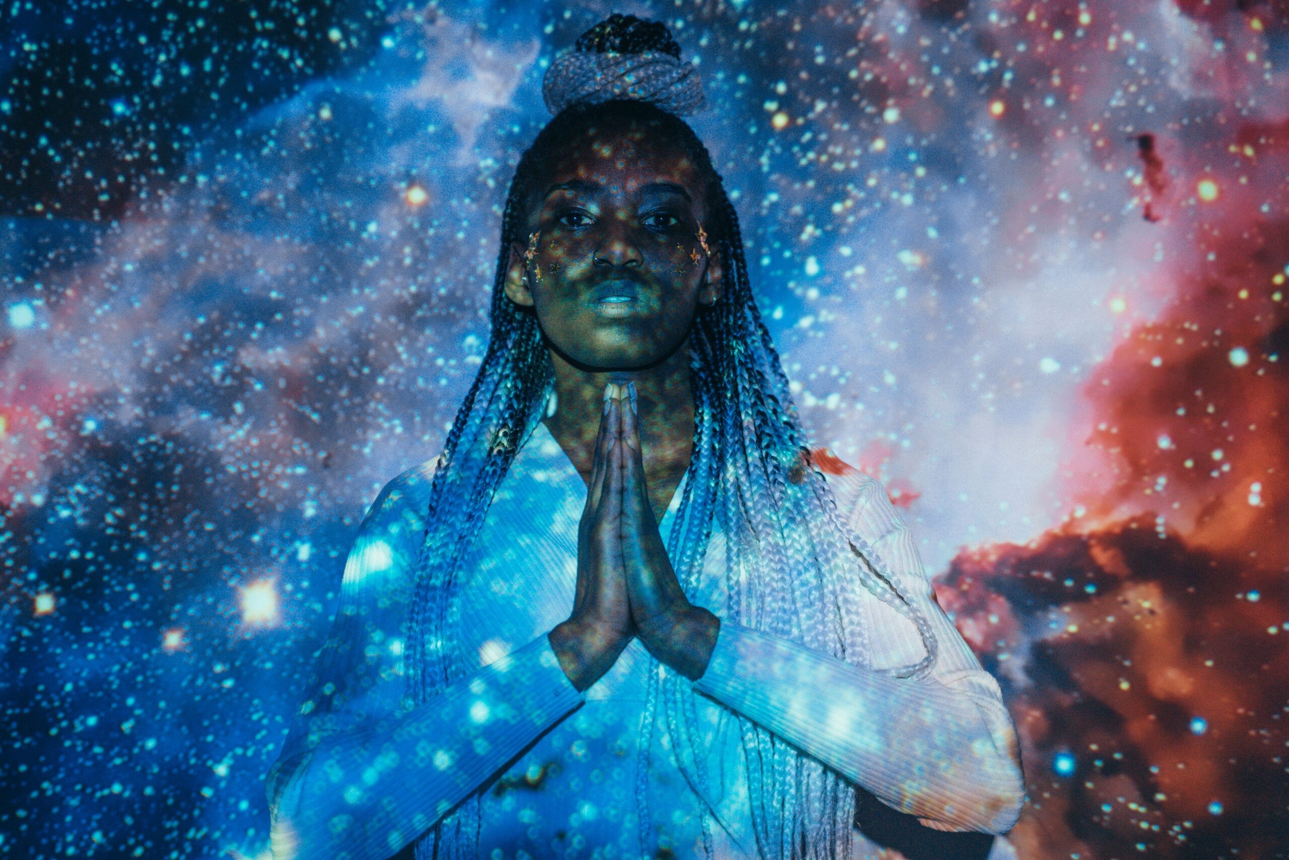 A woman with braided hair poses against a vivid galaxy projection with hands together.